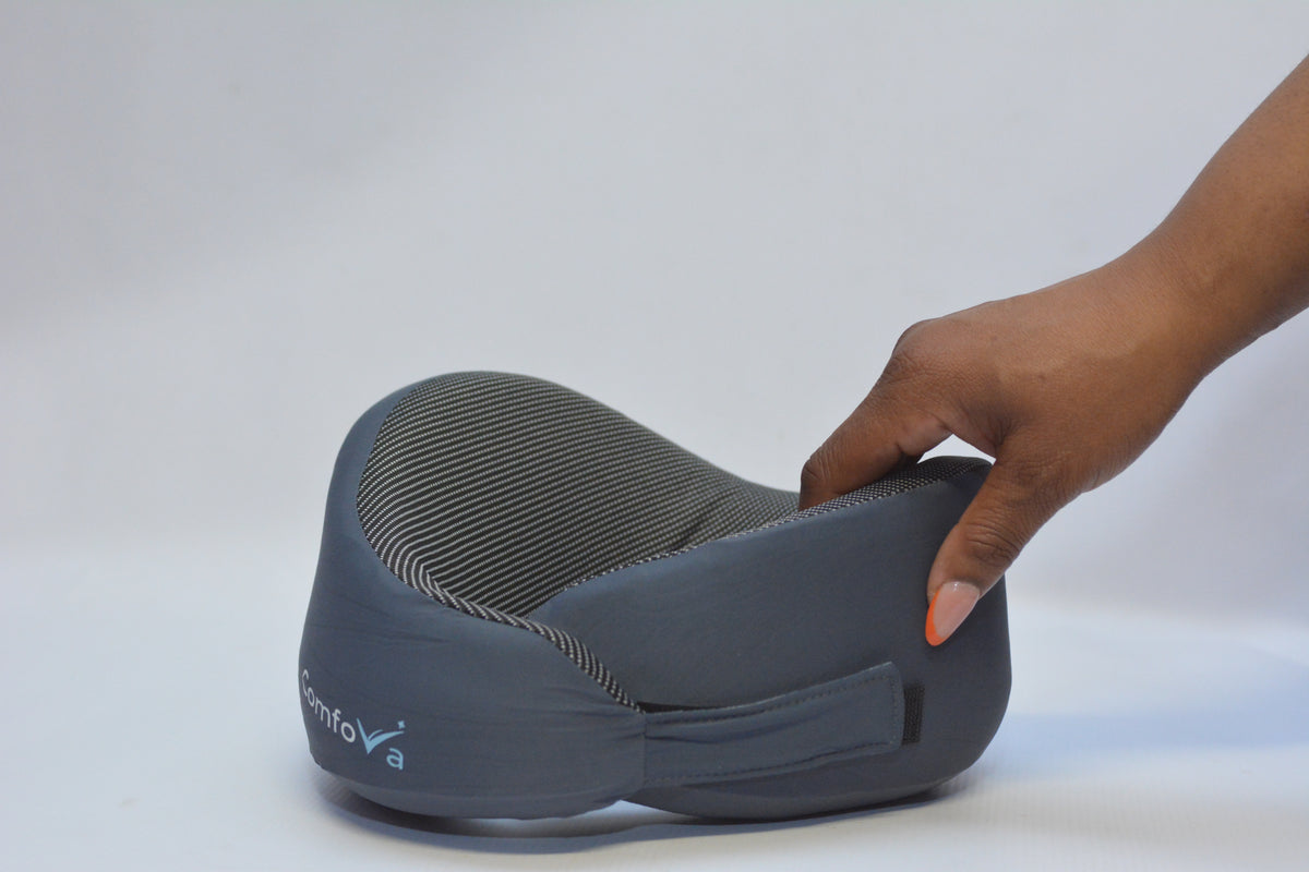 A dark gray contoured neck pillow with a textured striped interior, shown with a hand pressing down on the side to demonstrate its soft, flexible support.