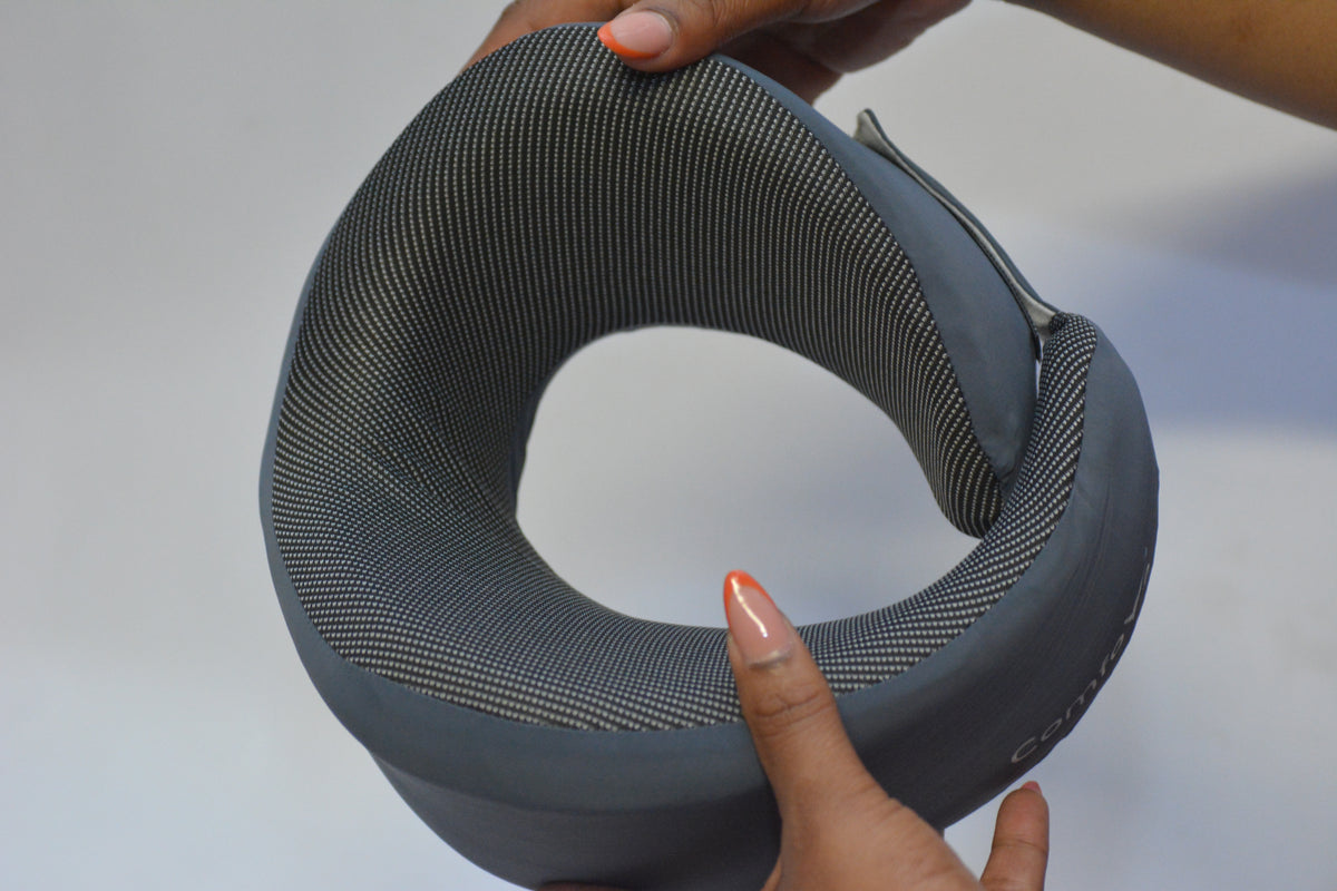 A gray contoured neck pillow with a textured mesh-like fabric, held by hands against a plain light gray background.