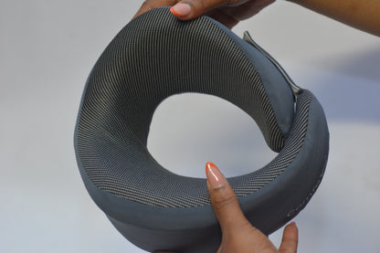 A gray contoured neck pillow with a textured mesh-like fabric, held by hands against a plain light gray background.
