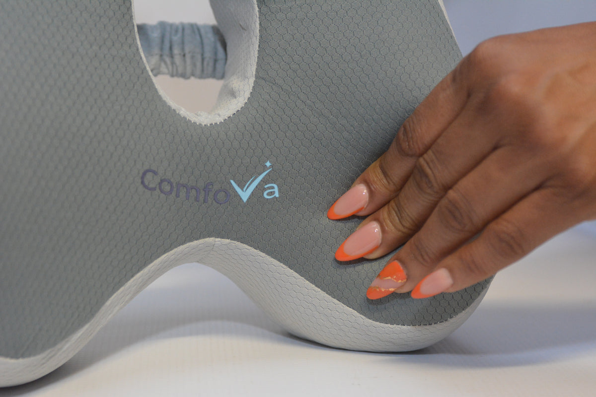 Close-up of a gray textured cervical pillow with the brand name 'ComfoVa' printed on it, being gently touched by a hand with orange manicured nails.