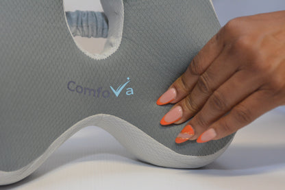Close-up of a gray textured cervical pillow with the brand name 'ComfoVa' printed on it, being gently touched by a hand with orange manicured nails.