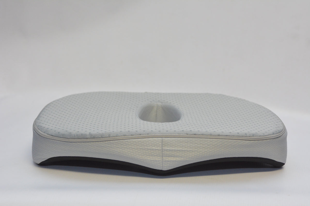 A light gray contoured lumbar support pillow with a central cutout, featuring a textured top surface and a black base, photographed on a seamless light gray background.