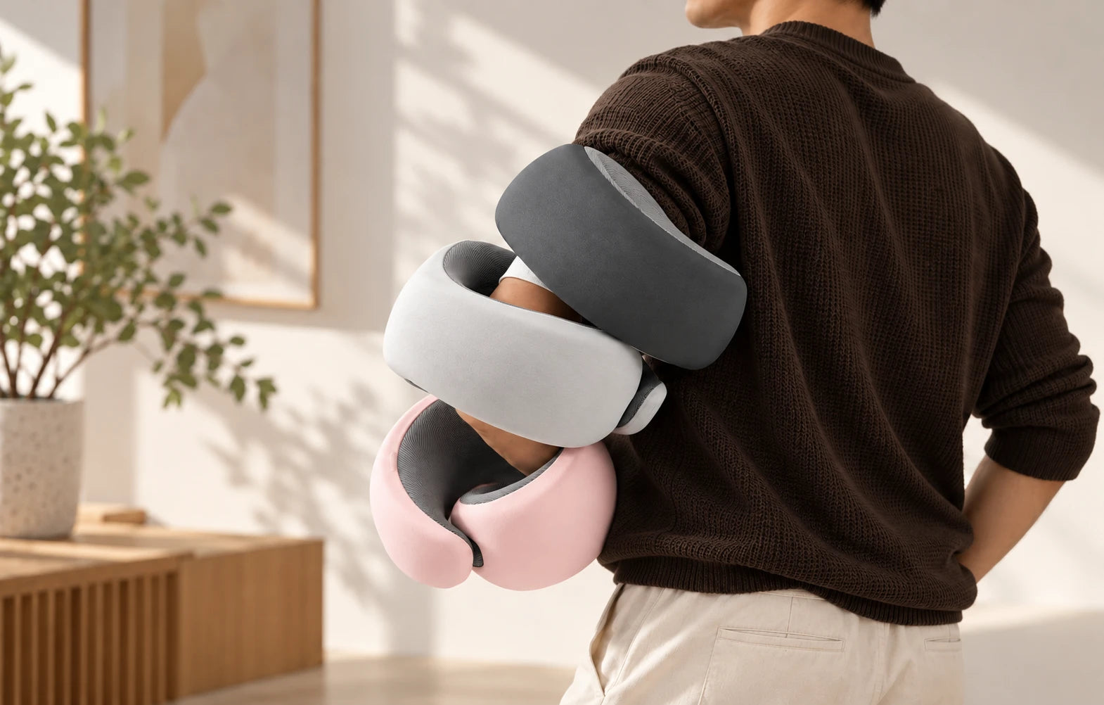 Three ComfoVa 360° travel pillows in Dark Grey, Light Grey and Light Pink, carried over a person's arm